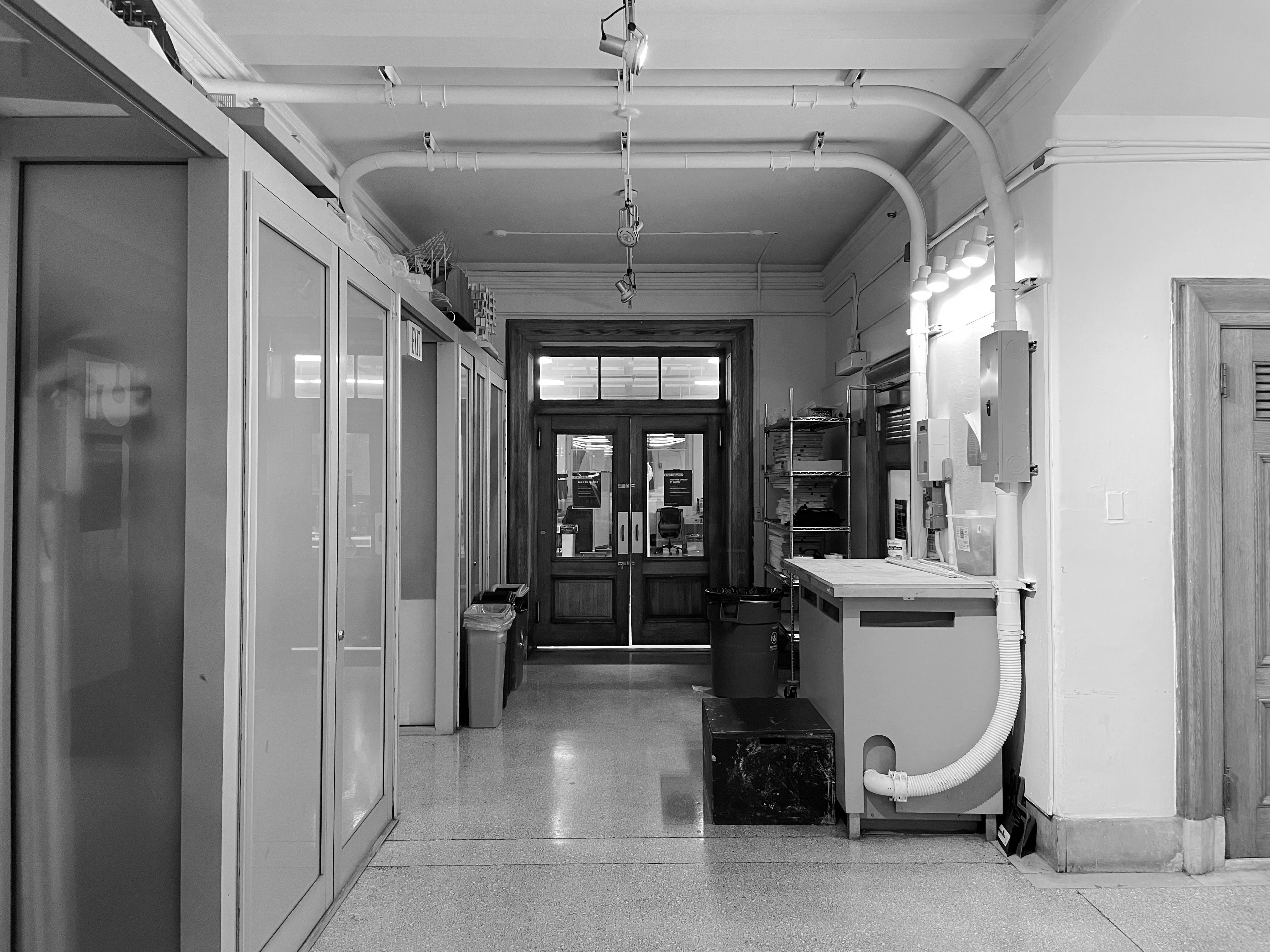 Black and white photo of an empty industrial hallway with pipes and cabinets.