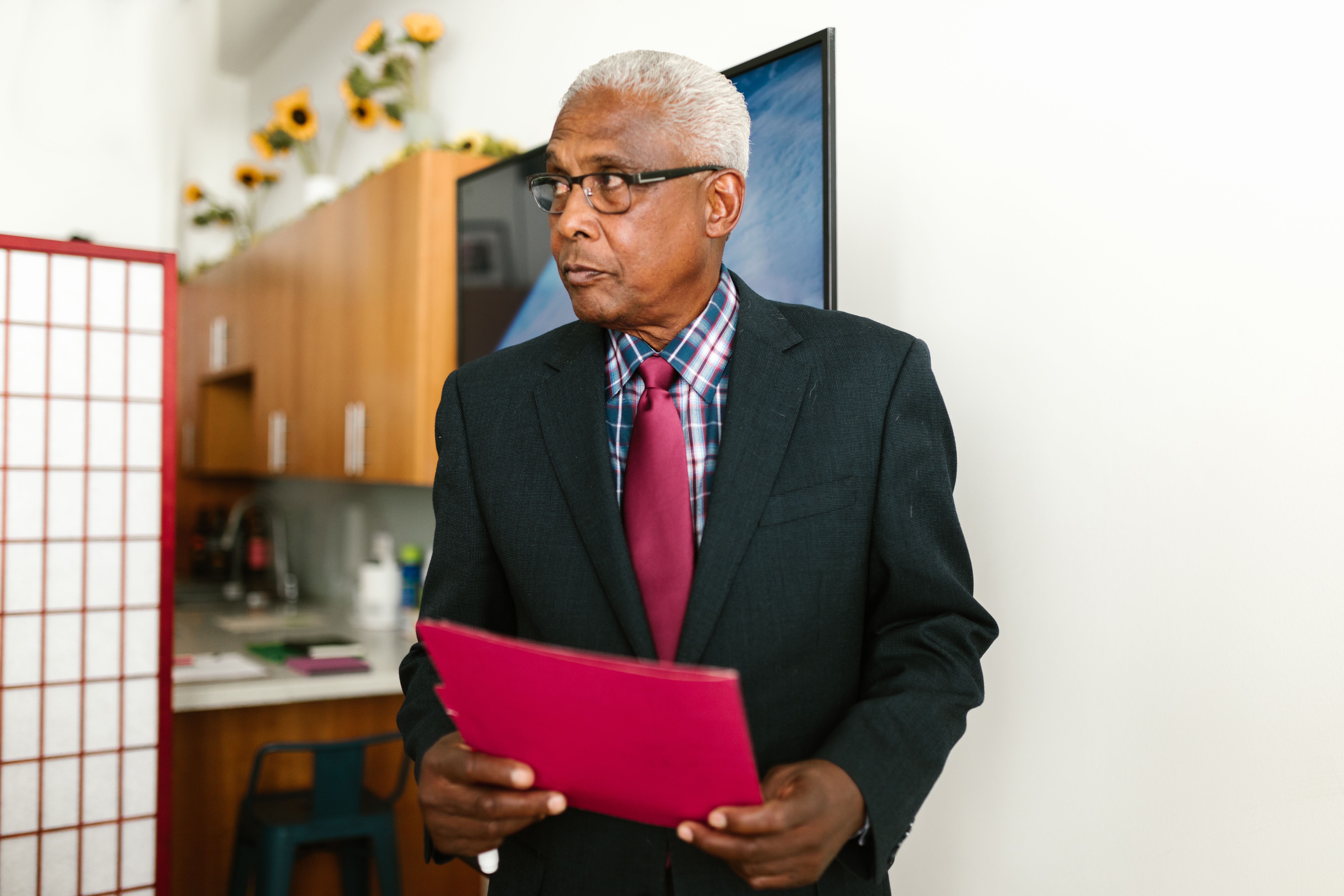 Senior businessman in suit holding a red folder, standing confidently in a modern office setting.