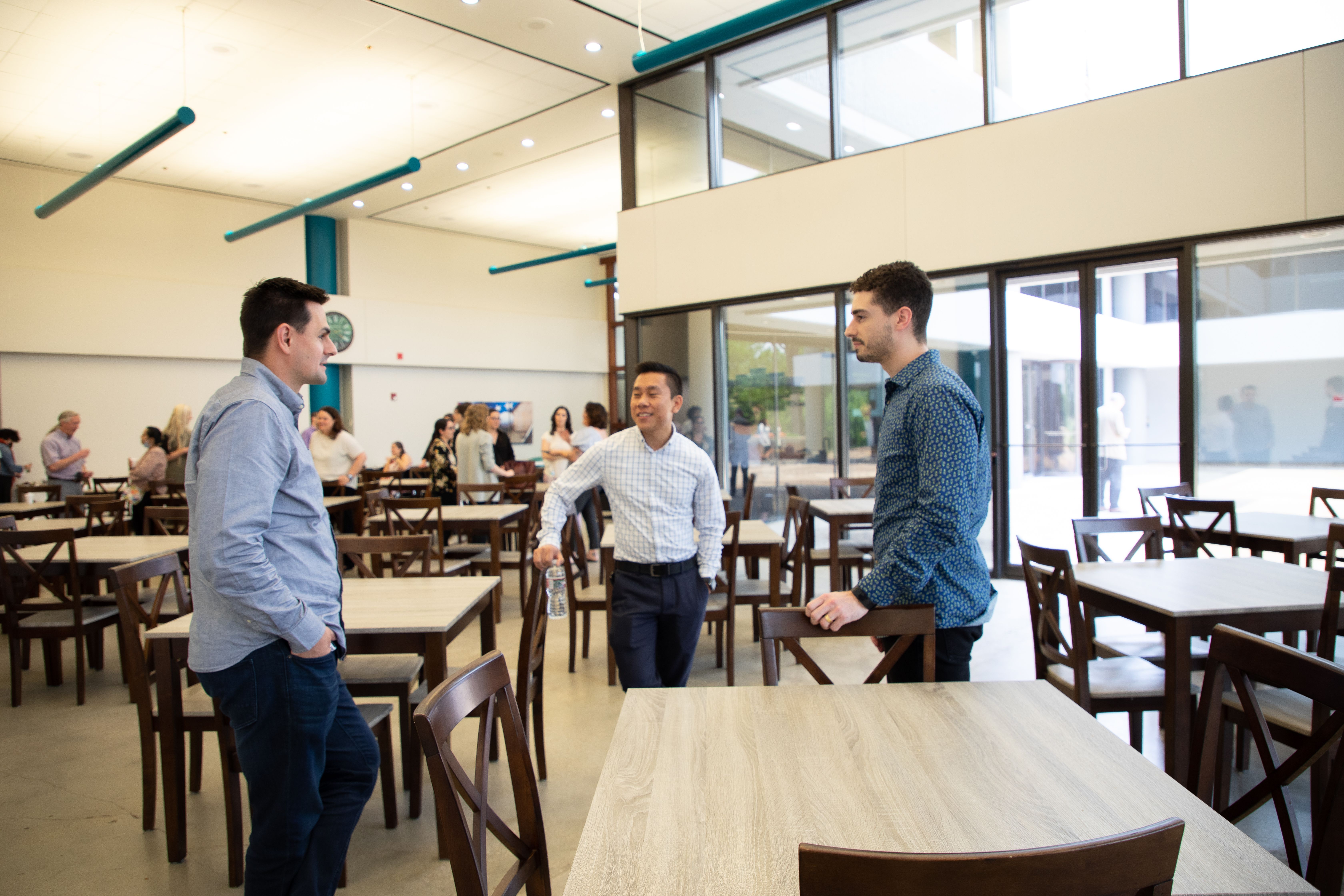 Three men engage in conversation in a modern, spacious cafeteria setting with chairs and tables.
