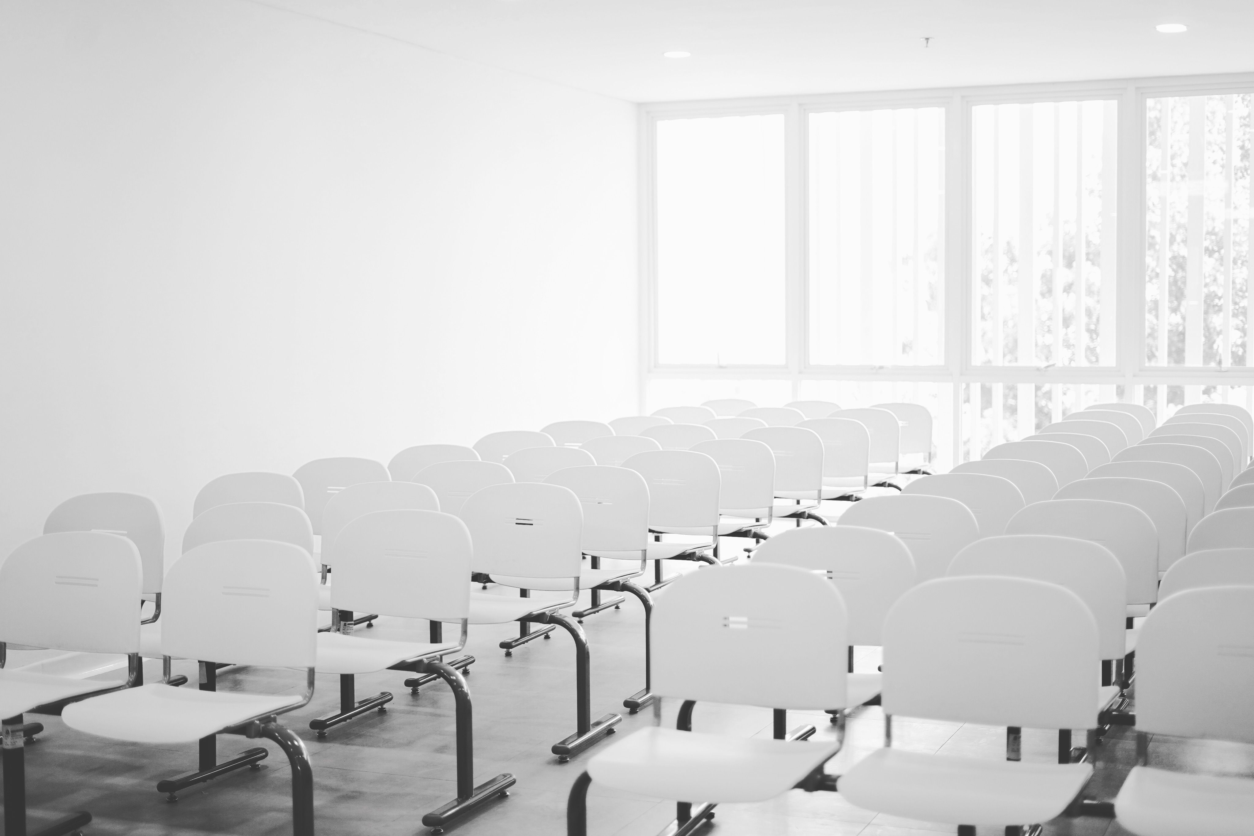 A bright, vacant conference room featuring rows of white chairs and natural light streaming through large windows.